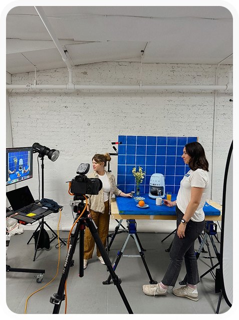 Two women setting up a product photo shoot with a blue tiled background, a coffee machine, flowers, and fruit on a table, with camera and lighting equipment around.